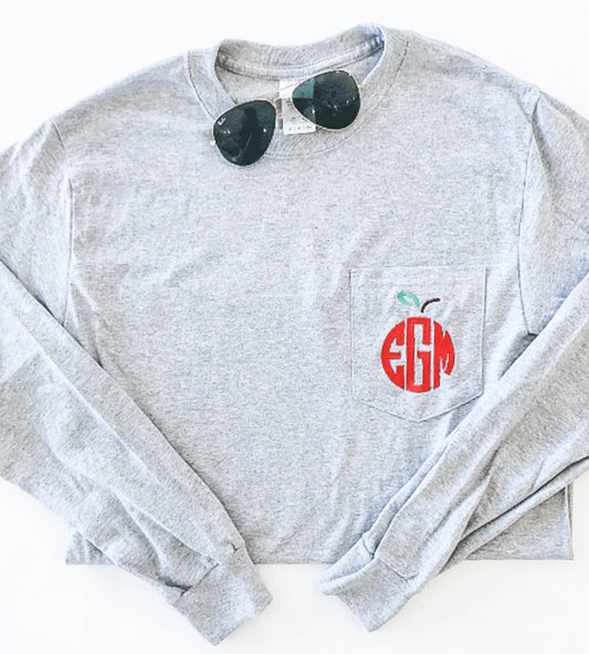 Red apple monogrammed embroidery on the pocket of a light gray long sleeved pocket top