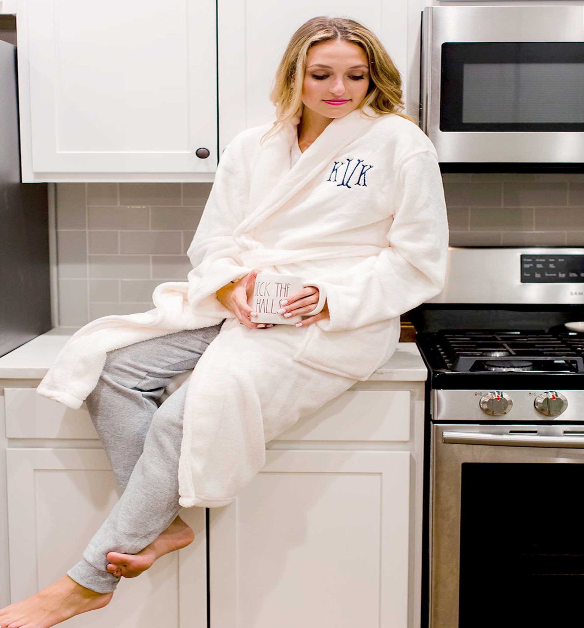 woman sitting on kitchen counter, holding a mug and  wearing a plush ivory robe with custom embroidered initials on the left chest.