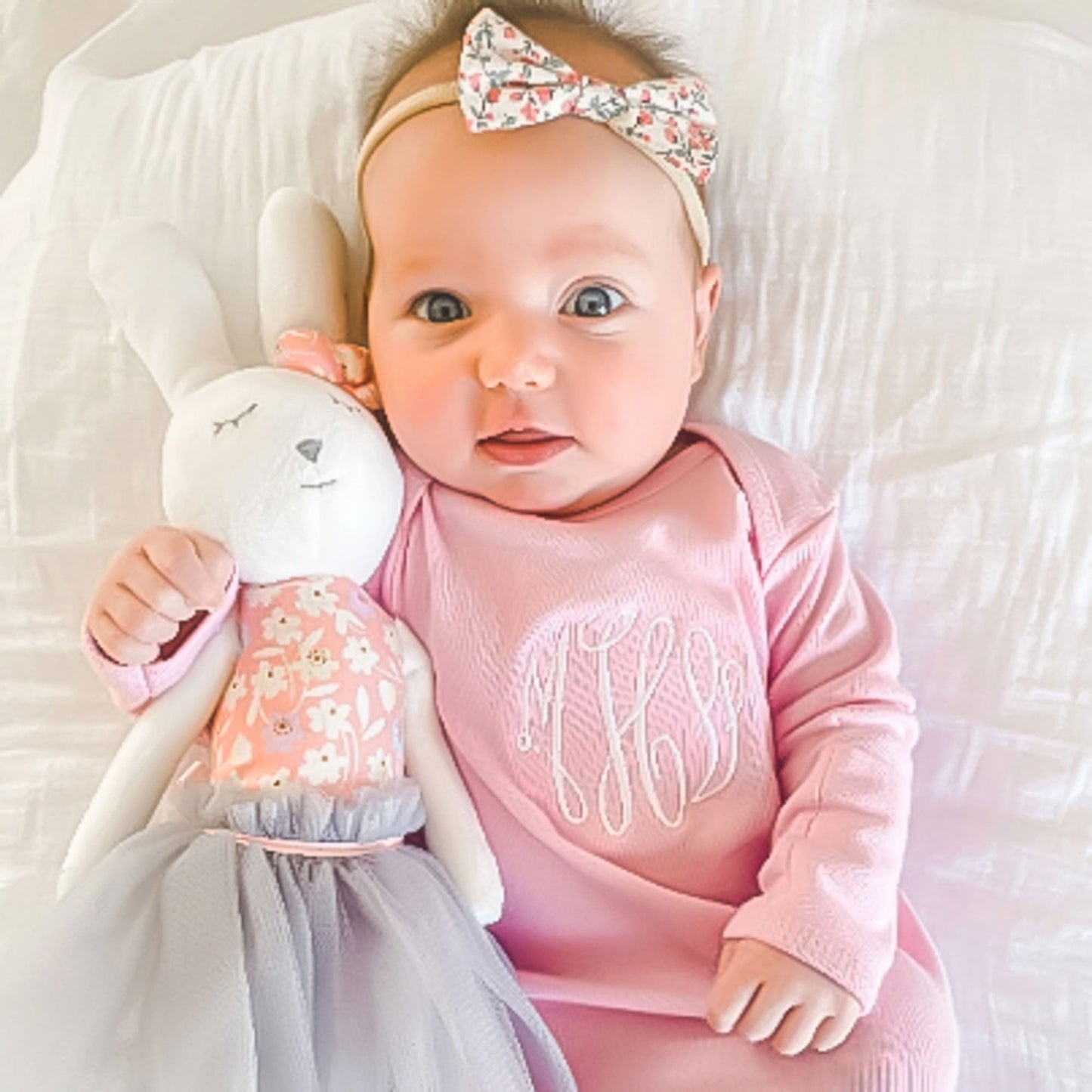 baby girl snuggling with toy bunny wearing pale pink layette with monogram embroidered on chest