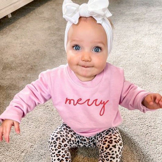 little girl with large bow on head wearing pink crewneck with word merry embroidered across chest in raspberry thread