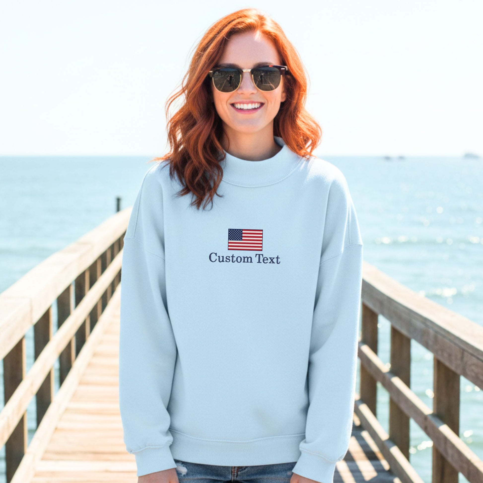Person wearing a light blue sweatshirt with an American flag and 'Custom Text' on a wooden pier by the ocean.