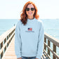 woman standing on a pier by the beach wearing a blue mockneck sweatshirt with custom embroidered usa flag and text design on the center chest