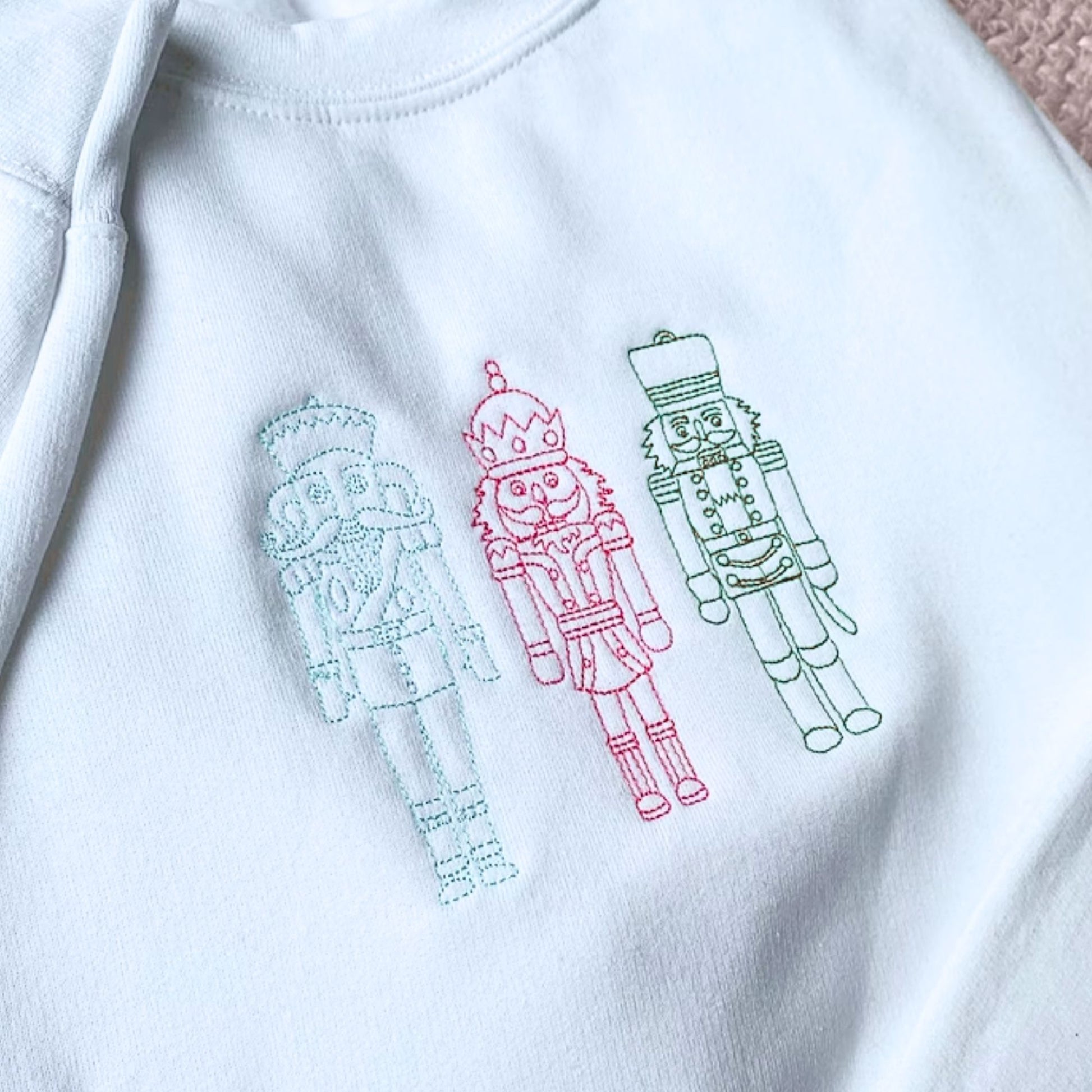 closeup of a  white crewneck sweatshirt with embroidered stitched nutcrackers across the chest in sky blue, pink, and kelly green