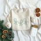 toddler heather oat crewneck sweatshirt with 3 outlined candy canes wrapped in bows embroidered across the chest in kelly green thread.