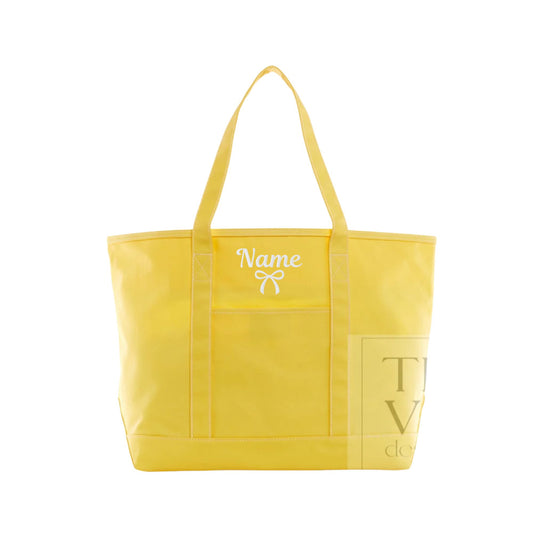 A bright yellow canvas tote bag with two shoulder straps, featuring a white embroidered 'Name' text and bow design on the front pocket, and a gold-toned printed logo on the lower right front panel.