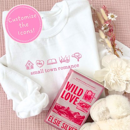 White crewneck sweatshirt with pink text and romance bookish icons, 'Wild Love' book by Elsie Silver, and decorative flowers on a pink background.