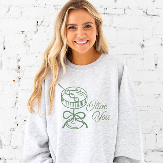 Woman wearing a gray sweatshirt with 'Olive You' design against a white brick wall.