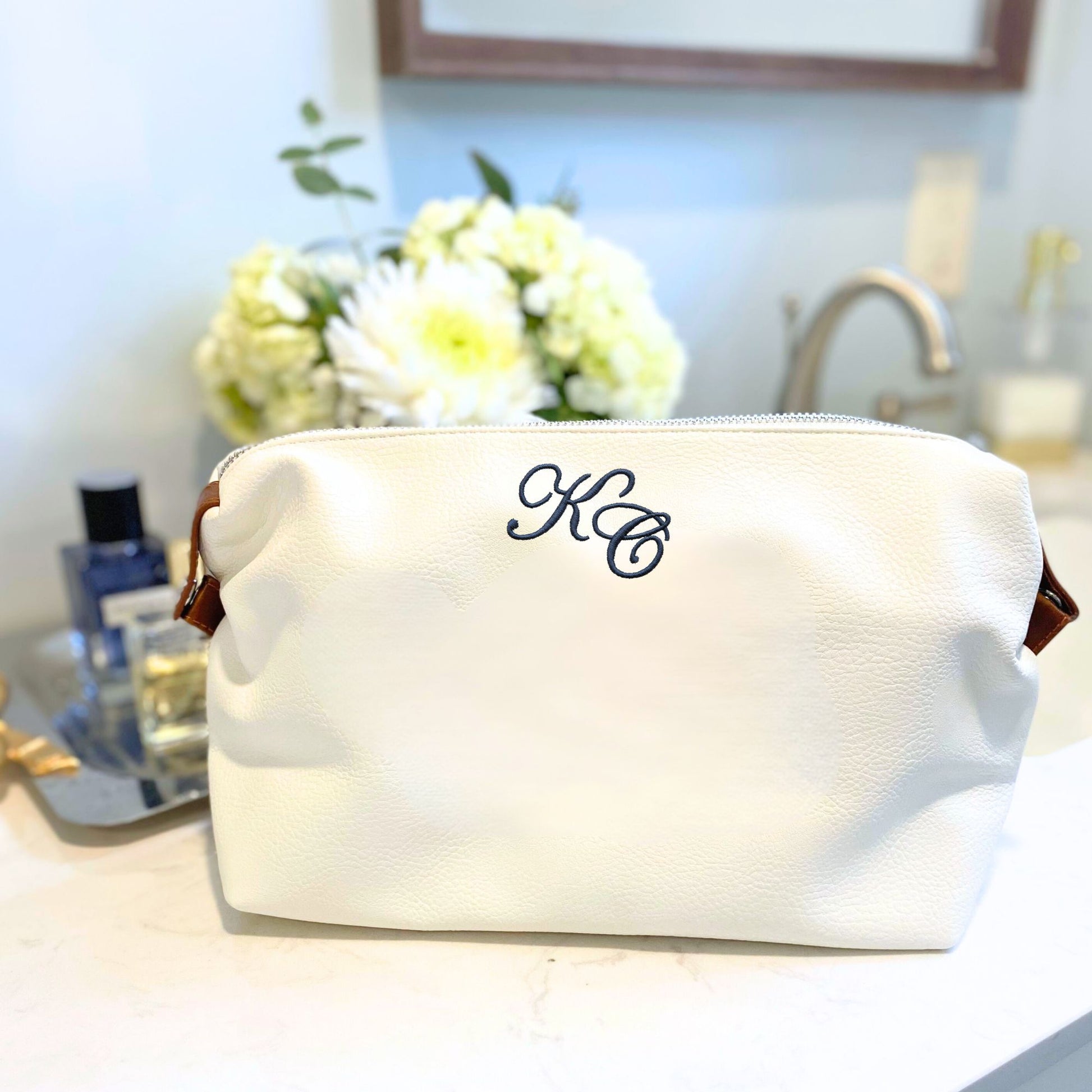 White cosmetic bag with 'KC' monogram on a bathroom counter with flowers and toiletries.