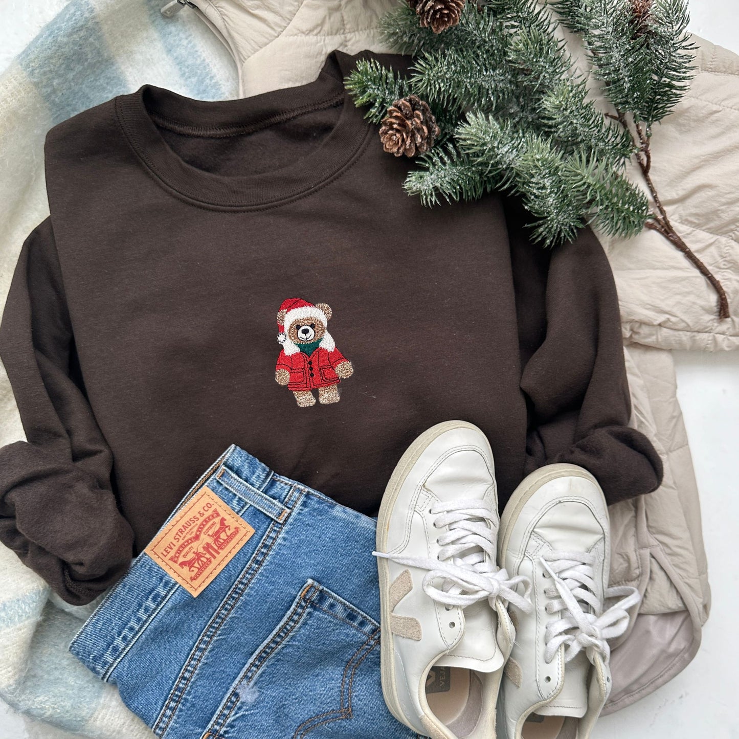 Brown sweatshirt with a teddy bear design, Levi's jeans, white sneakers, and Christmas decorations on a soft surface.