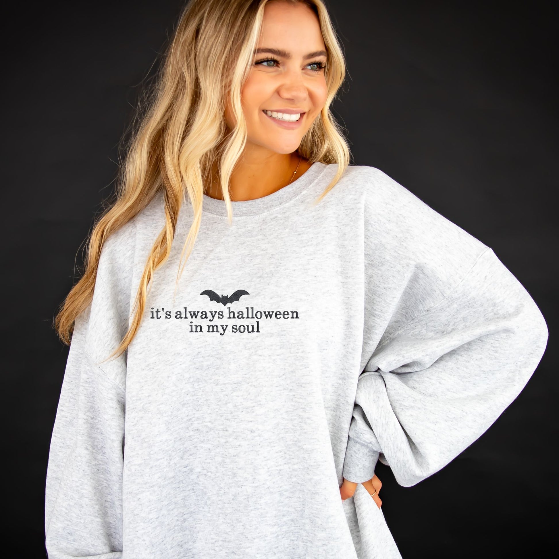 young woman wearing an oversized ash crewneck sweatshirt with embroidered its always halloween in my soul and bat design across the chest in black thread 
