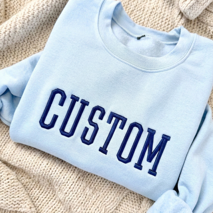 Light blue sweatshirt with 'CUSTOM' embroidered in dark blue on a textured beige surface