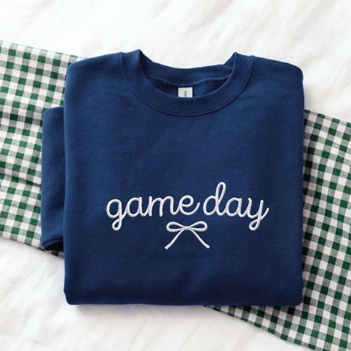 A folded navy blue sweatshirt with 'game day' and a bow graphic in white embroidery, displayed on a white textured surface with a green and white gingham blanket.