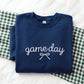 A folded navy blue sweatshirt with 'game day' and a bow graphic in white embroidery, displayed on a white textured surface with a green and white gingham blanket.