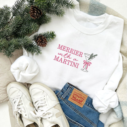 flat lay of a white crewneck sweatshirt with embroidered merrier with a martini text in pink thread and martini glass design with bow and pine sprig across the chest