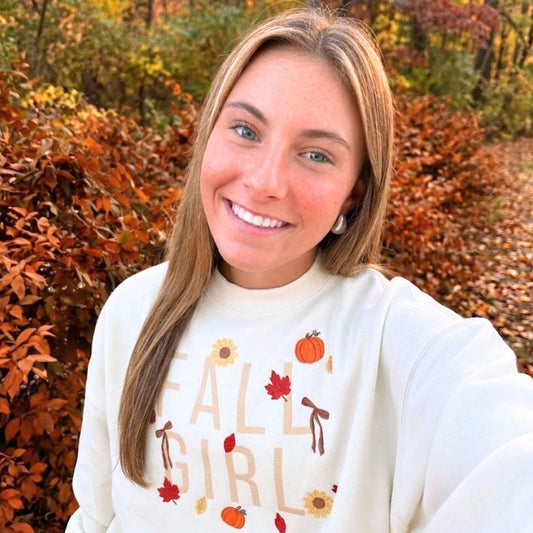 A smiling woman taking a selfie outdoors in autumn foliage, wearing a cream sweatshirt with 'FALL GIRL' and fall-themed graphics.