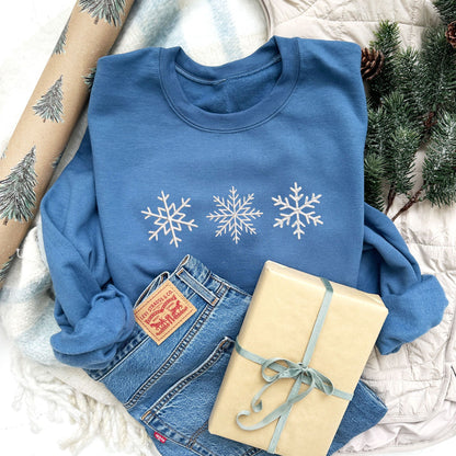 Christmas styled flat lay photo an indigo crewneck sweatshirt with3 embroidered snowflakes across the chest