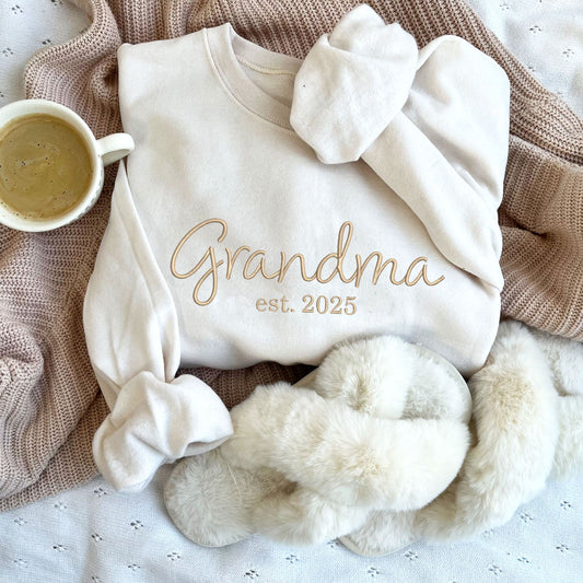 Flat lay of a sweet cream crewneck sweatshirt styled with coffee and slippers. On the center chest of the crewneck is embroidered Grandma est2025 in camel thread.