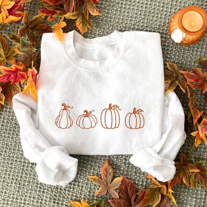 flatlay of a white crewneck sweatshirt with 4 different pumpkin outlines embroidered across the chest in pumpkin spice thread