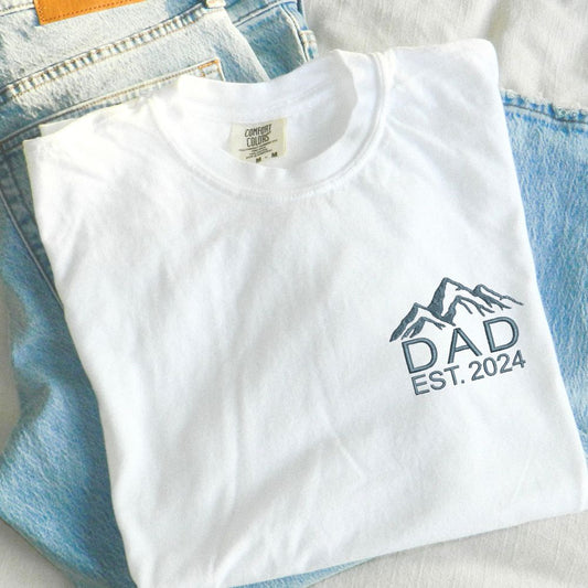 a white t-shirt with custom mountain icon and dad/custom year embroidered design on the left chest in French blue thread