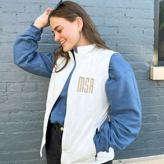 young woman wearing a white softshell vest with MSA initials embroidered on the left chest in camel thread.