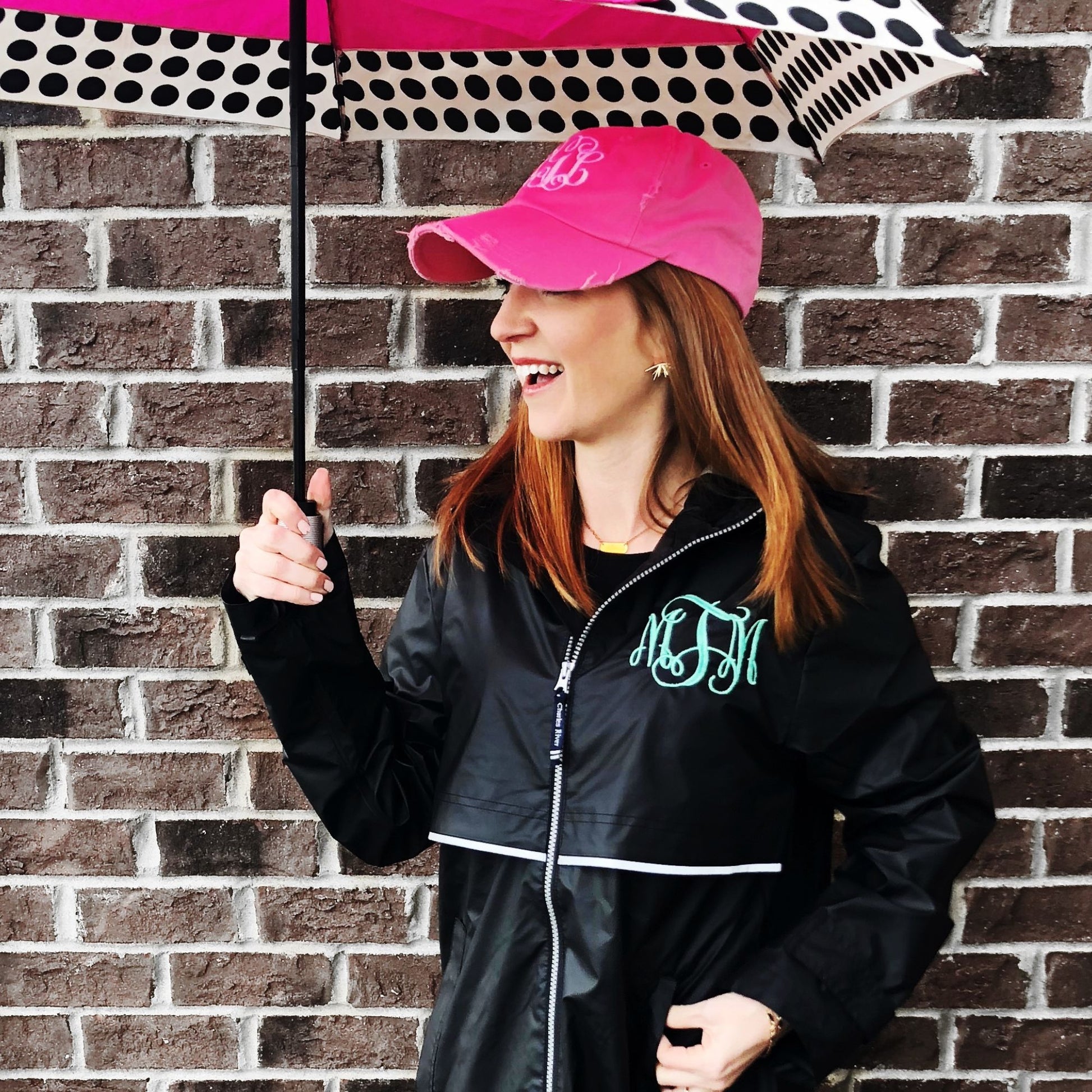 woman standing outside holding an umbrella wearing a black hooded full zip rain coat with custom monogram embroidered on the left chest in aqua thread.