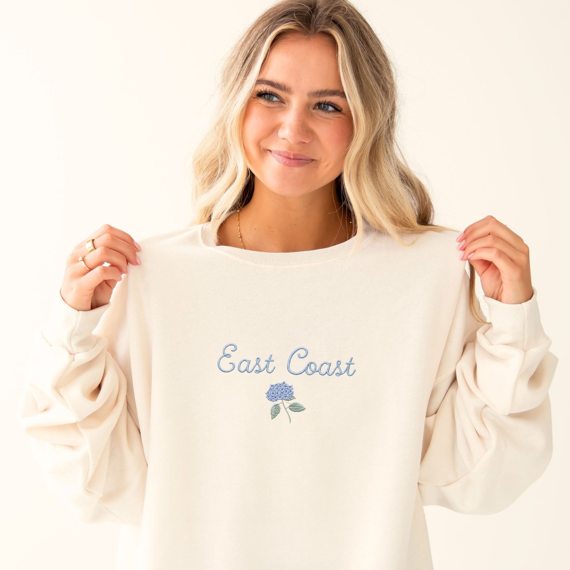 Woman wearing a sweet cream sweatshirt with 'East Coast' text against a white brick wall.