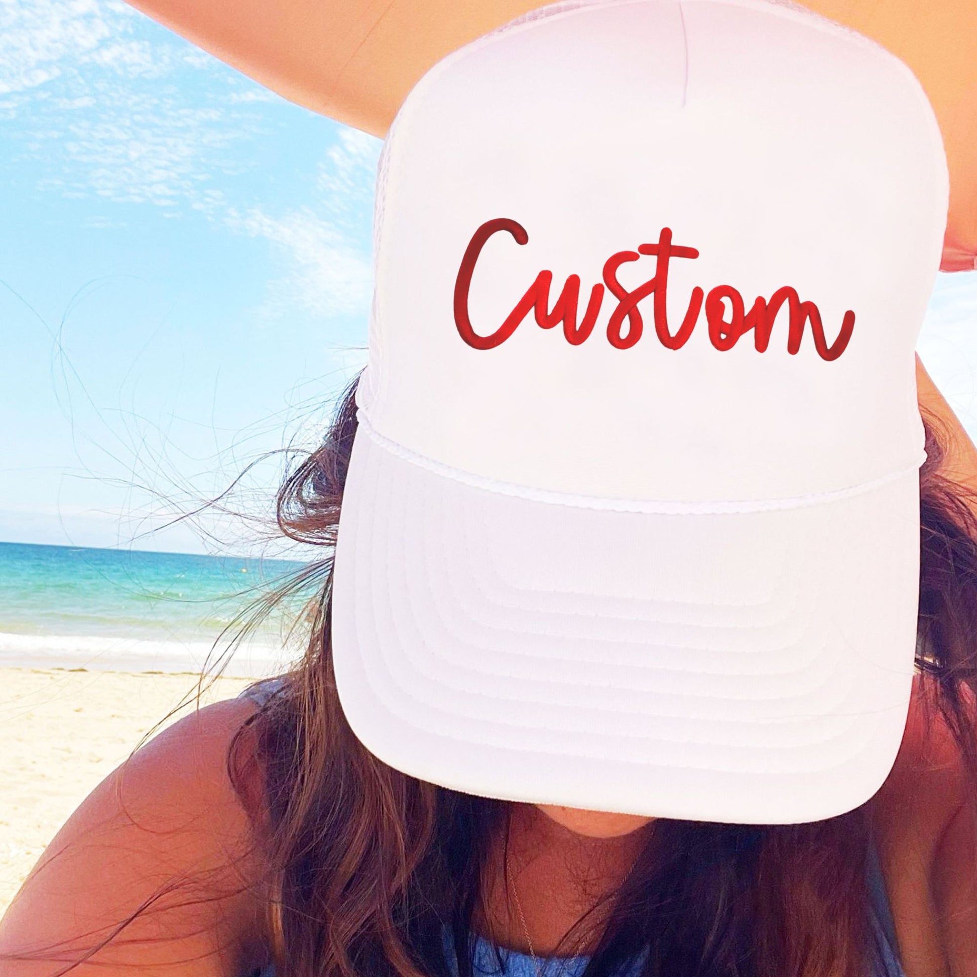 Person wearing a white cap with 'Custom' embroidered in a 3d puff script font in red on a beach.