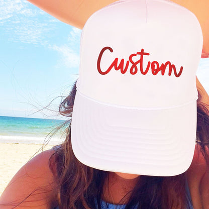 Person wearing a white cap with 'Custom' embroidered in a 3d puff script font in red on a beach.