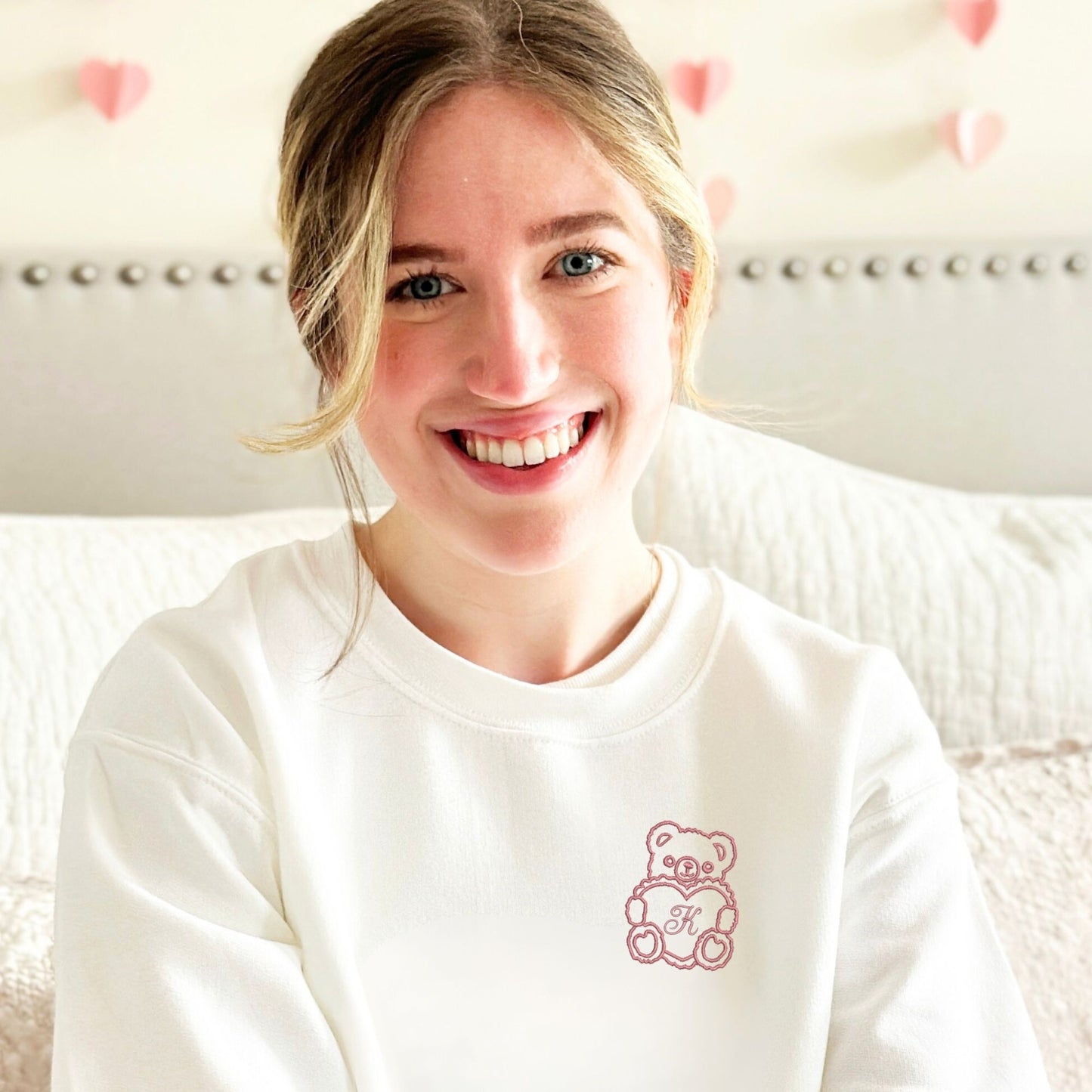 girl wearing a white crewneck sweatshirt with a design of an outlined teddy bear holding a heart with an initial in the center embroidered on the left chest in mauve thread