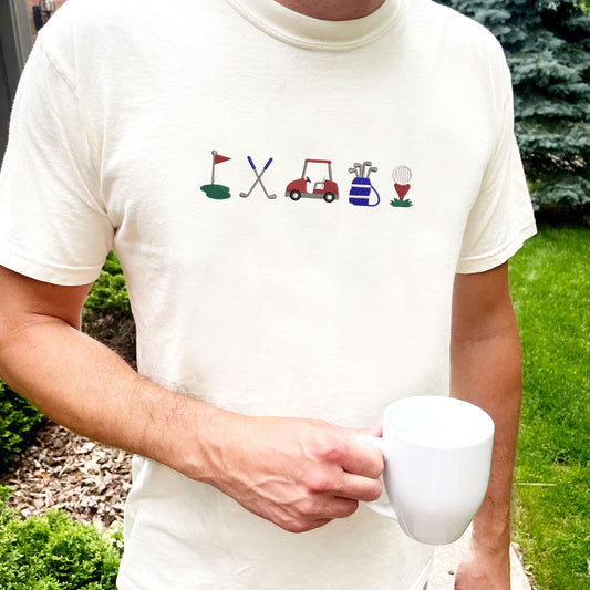 man wearing an ivory comfort colors t-shirt with custom embroidered golf icons - golf flag, golf poles, golf cart, golf bag, and ball with tee in shades of blue, green, and red