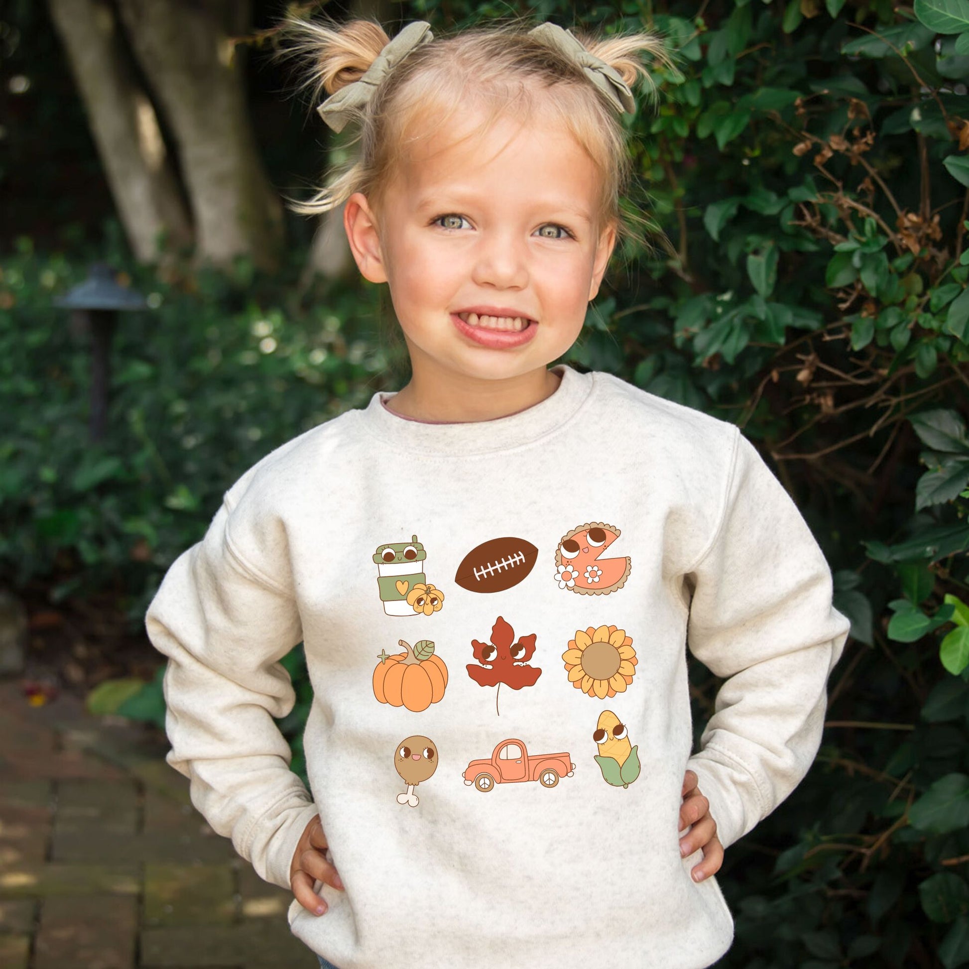 Little girl wearing a heather oatmeal crewneck sweatshirt with cute fall icons like a football, pumpkin, leaves, turkey leg, truck, and sunflower