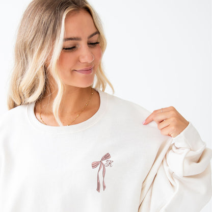 girl wearing a sweet cream sweatshirt with a ribbon bow and single initial embroidered design on the left chest in mauve thread