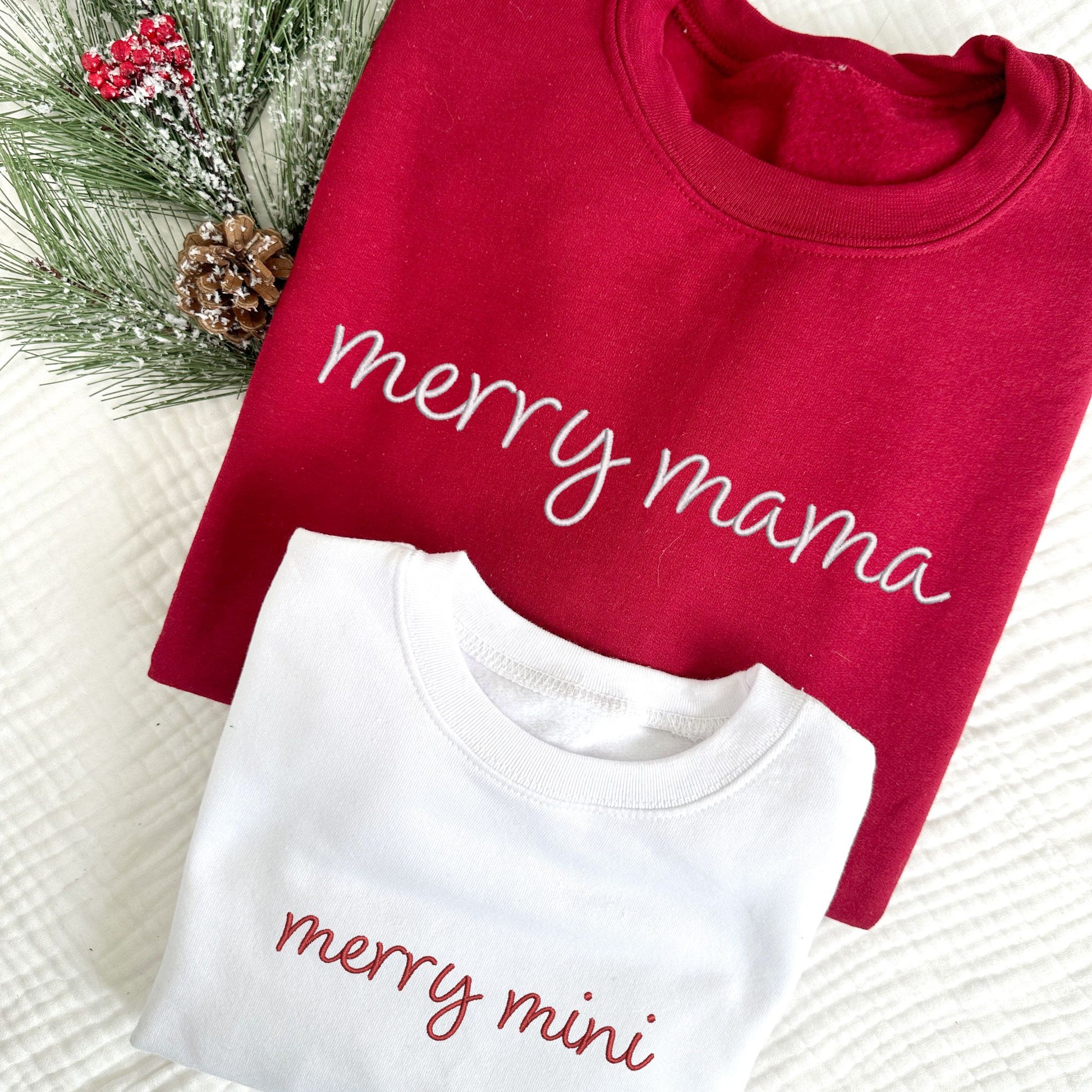 cardinal red crewneck sweatshirt with embroidered merry mama in white thread and a toddler white crewneck with merry mini embroidered in red thread across the chest