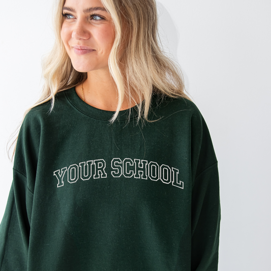 young woman wearing a forest green crewneck sweatshirt with custom athletic block outline embroidered font across the chest 