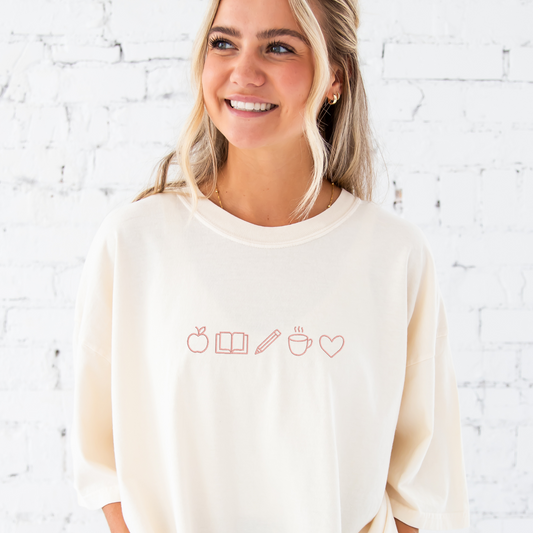 young woman wearing a large ivory tee with embroidered teacher icons in coral pink thread across the chest