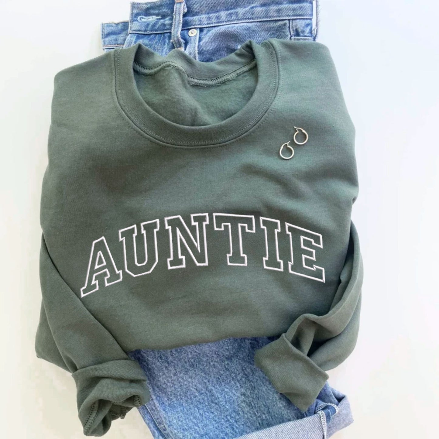 flay lay image of jeans and a olive green crewneck sweatshirt with embroidered AUNTIE in all caps athletic block font across the chest in white thread.