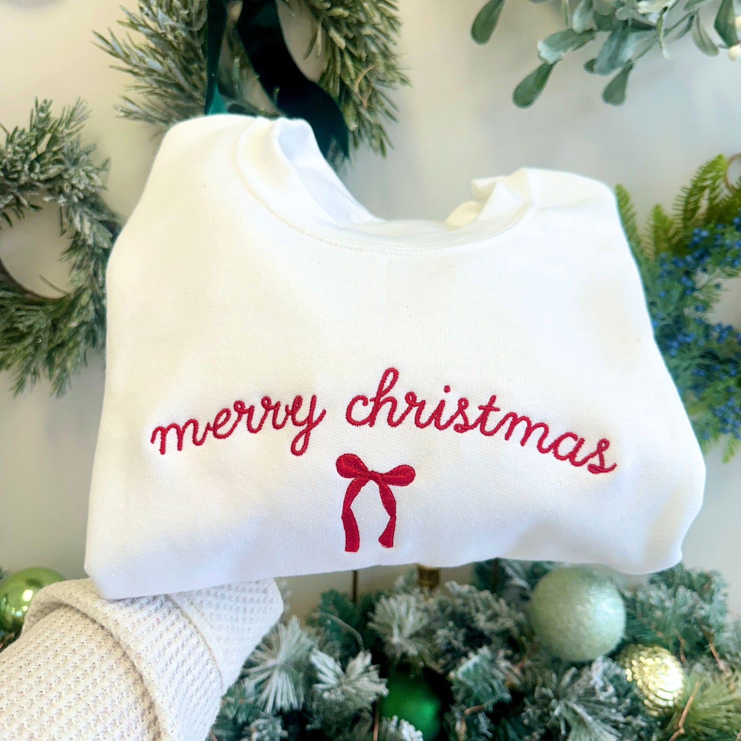White sweatshirt with 'merry christmas' text and red bow held in front of a Christmas tree.