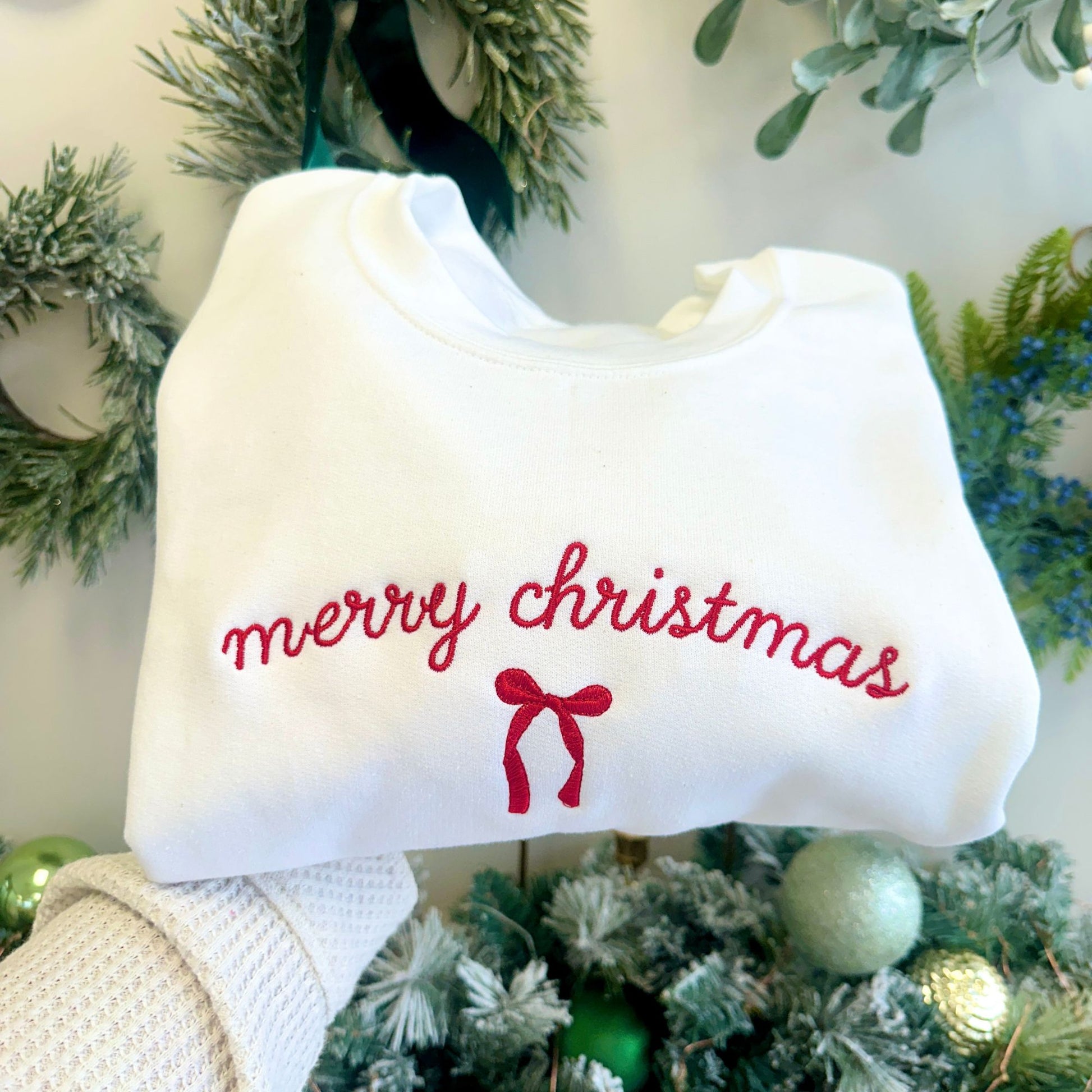 White sweatshirt with 'merry christmas' text and red bow held in front of a Christmas tree.