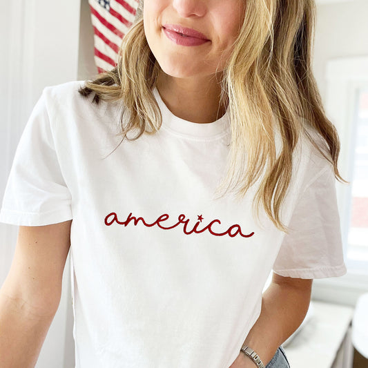 girl wearing a white comfort colors tee with a cute america script embroidered design across the chest