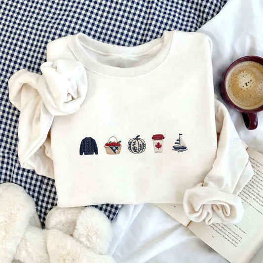 A folded cream-colored sweatshirt with a row of small embroidered icons across the chest, styled in a cozy flat-lay arrangement with a gingham blanket, a coffee cup, an open book, and soft white fabric.