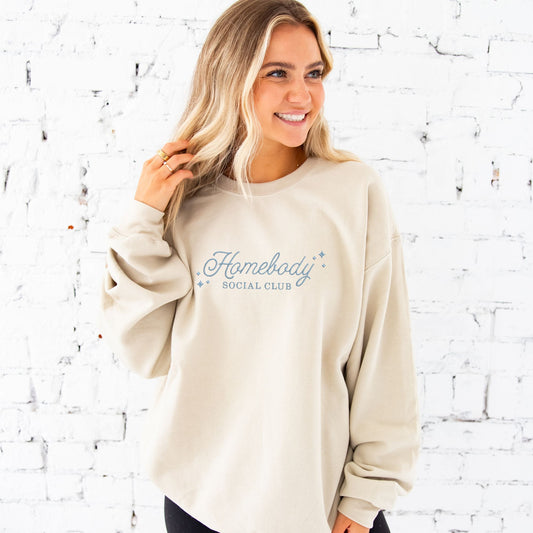 young woman modeling a sand crewneck sweatshirt with embroidered homebody social club design in baby blue thread c