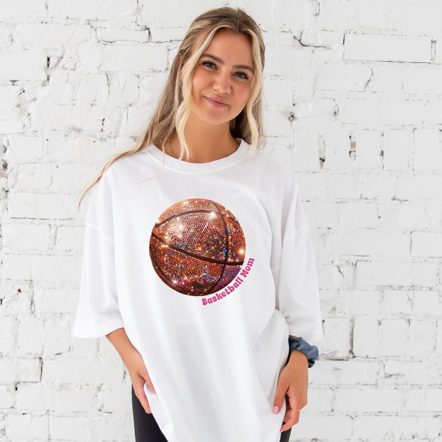 young woman wearing a white comfort colors t-shirt with large glitter basketball print with basketball mom text wrapping around the ball in pink ink