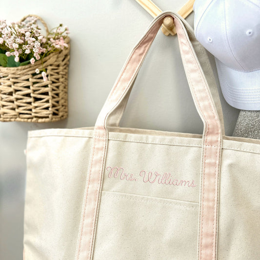Canvas tote bag with a powder pink trim and custom name embroidery hanging on a natural wood hook next to a white baseball hat and a small wicker basket containing pale pink flowers