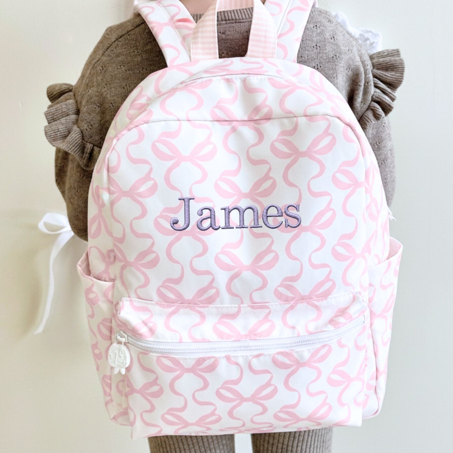 little girl wearing a white and pink bow patterned backpack with 'James' embroidered on it in smoky orchid thread