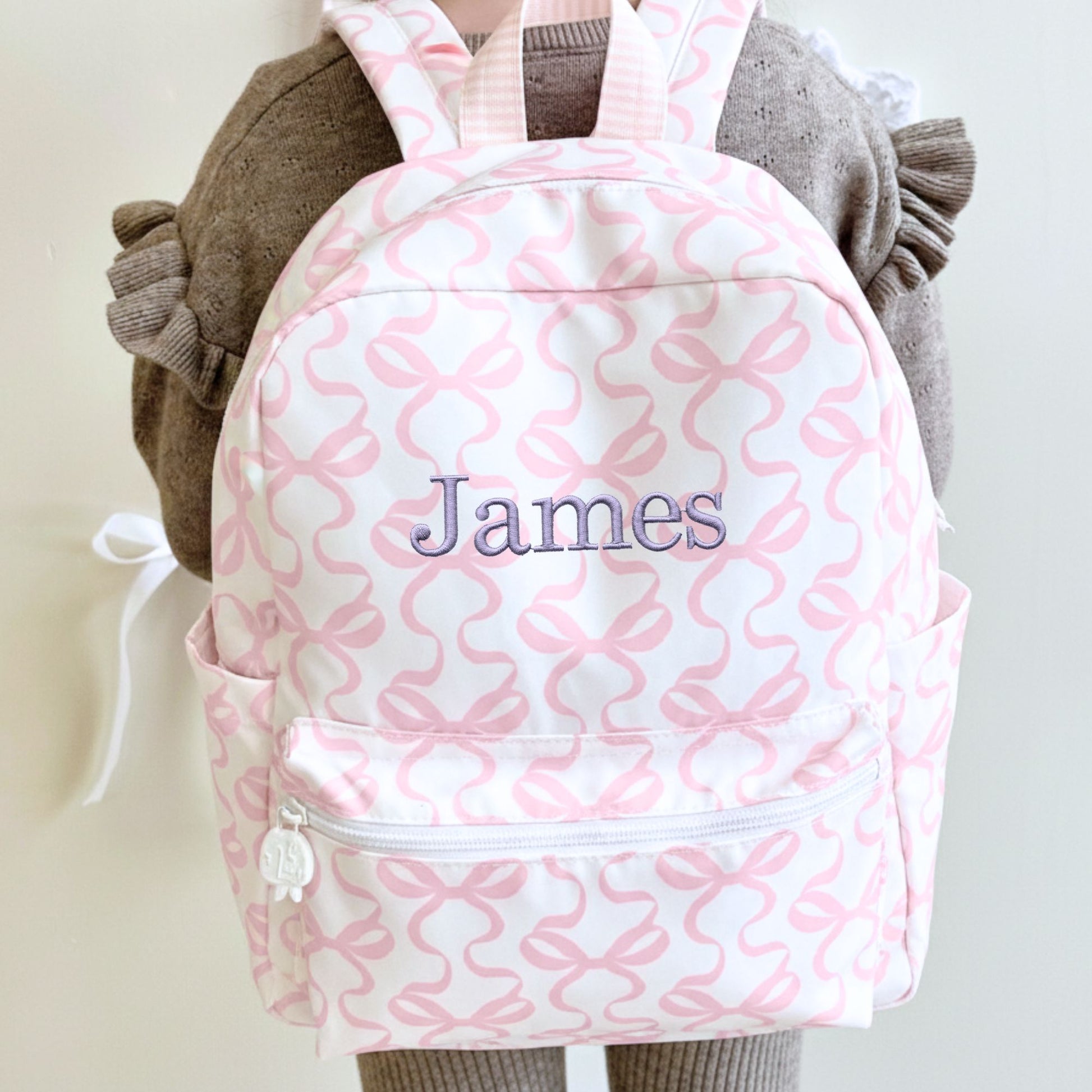 little girl wearing a white and pink bow patterned backpack with 'James' embroidered on it in smoky orchid thread