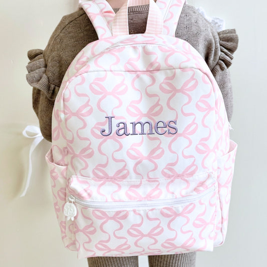 little girl wearing a white and pink bow patterned backpack with 'James' embroidered on it in smoky orchid thread