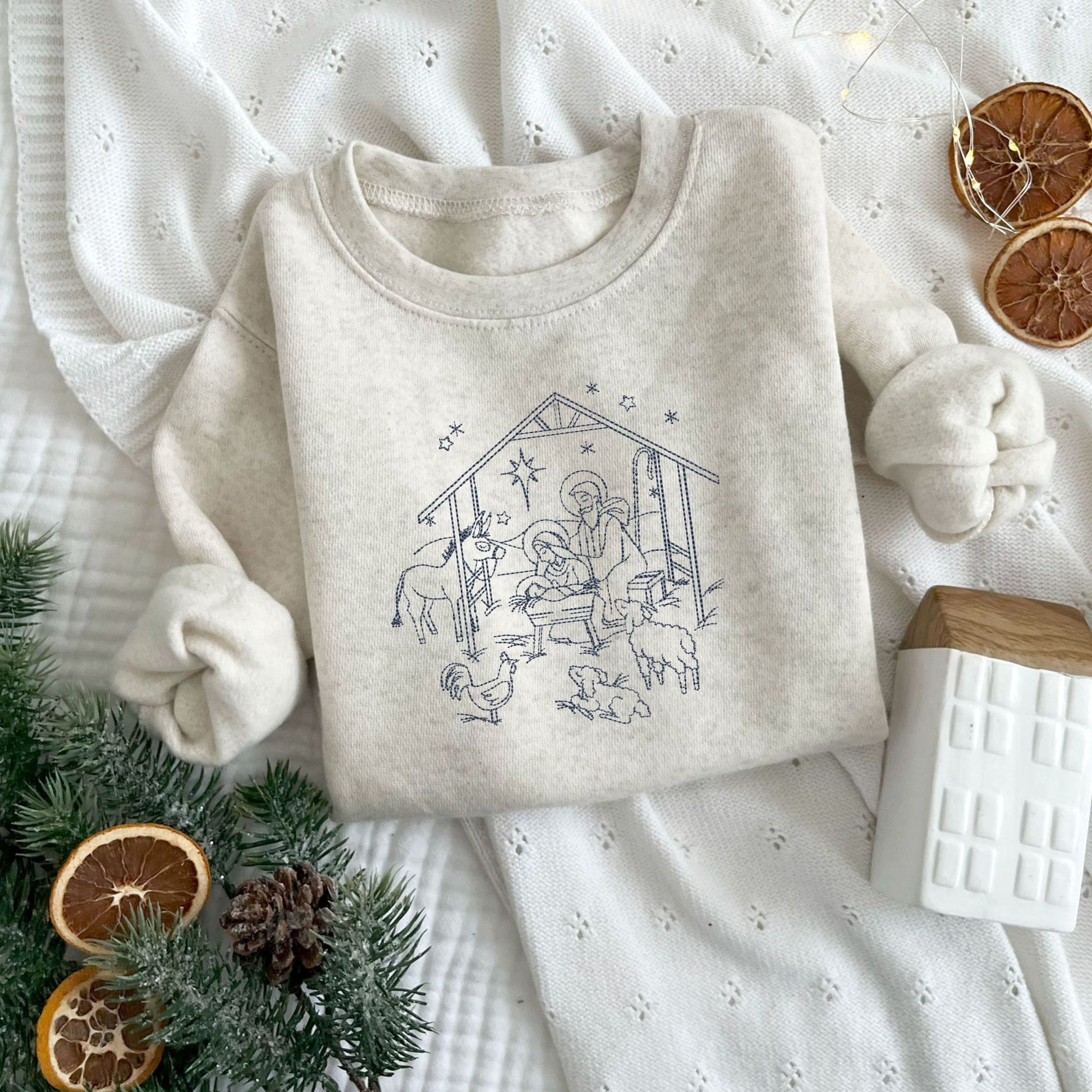 Christmas styled flat lay of a toddler heather oatmeal crewneck with embroidered stitched nativity scene in navy thread