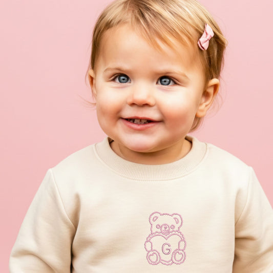 little girl wearing a natural crewneck sweatshirt with embroidered teddy bear heart initial design on the center chest in baby pink thread.