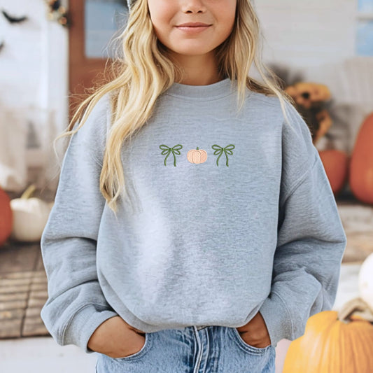 Little girl wearing a gray sweatshirt with pumpkin and bows embroidered design in pink apricot and olive green thread. She's standing on a festive fall decorated porch with pumpkins.