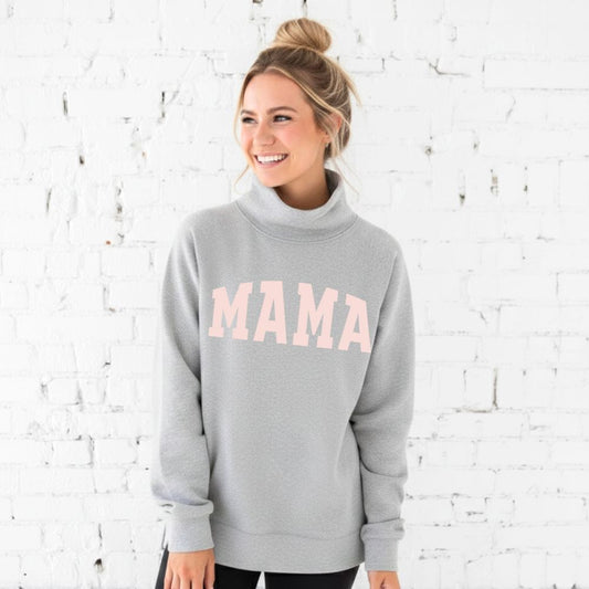 Woman wearing a gray sweatshirt with 'MAMA' printed on it against a white brick wall.